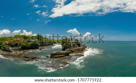 Panoramic picture of tanah lot in bali, indonesia, with the temple in the center, a part of the touristic area on the left side, the sea on the right side, and the sky in the background