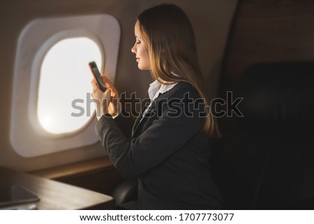 Adorable caucasian girl traveling by private airplane. Charming brown haired young woman sitting by aircraft window and taking pictures of sky on her smartphone. Flying first class.