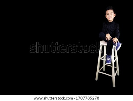 A five years old boy. A cute five year old boy studio portrait on black background. A boy wearing black jeans and black T-shirt. A child sitting on the white chair in studio.