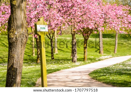 A sign on a wooden post reading the word "run" on a fitness trail by a sunny spring day in a public park with blossoming Japanese cherry trees in the background along a dirt path.