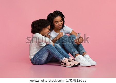 Smiling african girl and her mom sitting on floor with smartphone, watching video or cartoons from internet, free space