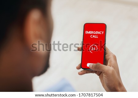 Emergency Call. View over shoulder of man holding phone with 24 hour support line on red screen over blurred background Royalty-Free Stock Photo #1707817504