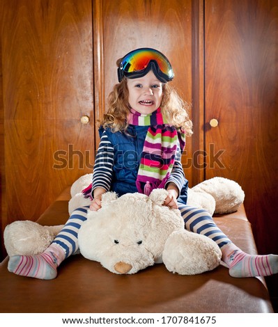 Little curly-haired girl in ski glasses sitting on a big teddy polar bear. Shot indoors.
