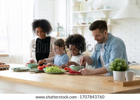 Happy young multiethnic family with little daughters have fun cooking at home together, smiling parents and small multiracial children prepare breakfast salad in kitchen, healthy lifestyle concept
