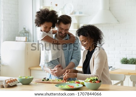 Happy young diverse parents have fun teach little biracial daughter cooking, overjoyed multiracial family with small girl child preparing food making healthy salad for breakfast in kitchen together Royalty-Free Stock Photo #1707844066