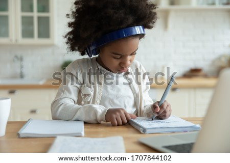 Smart little African American girl in headphones watch online video lesson at home, small biracial child in earphones have web video class on computer, study in kitchen, homeschooling concept