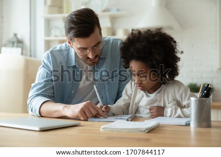 Caring Caucasian father help biracial little daughter with homework at home, loving European dad and small African American girl child study together in kitchen on quarantine, homeschooling concept Royalty-Free Stock Photo #1707844117