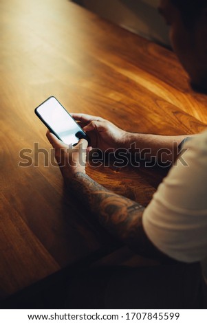  Mockup shot of man's hand with tattoos holding cell phone with blank screen on desk at home office.