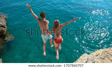 Joyful tourist couple decides to jump off a rocky cliff and dive into sea. Unrecognizable man holds his gorgeous girlfriend's hand while diving into the refreshing blue sea on a sunny summer day. Royalty-Free Stock Photo #1707860707
