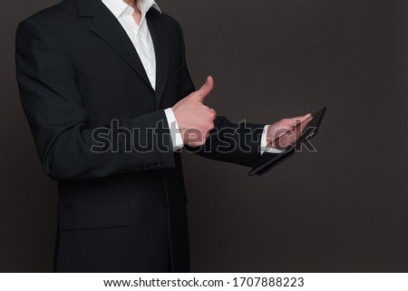 Businessman in black suit holding tablet pc in his hand closeup side view. Showing thumbs up sign into a camera or screen. Good deal, great offer