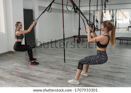 Young female people in sportswear performing TRX training in gym. Sporty girls doing exercise with elastic rope, full body, candidemotions, lifestyle concept.