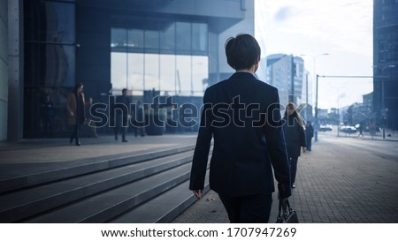 Businessman in a Suit Walking on the Street. Other Office People Commute to Work or Home. It's Early Morning or Late in the Evening. Back Shot. Urban City Lights in the Background. Royalty-Free Stock Photo #1707947269