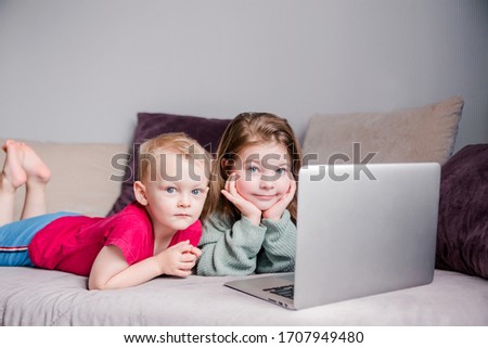 Two children a boy and a girl lie on a sofa in home clothes and watch videos on a laptop and look at camera for a moment. 
