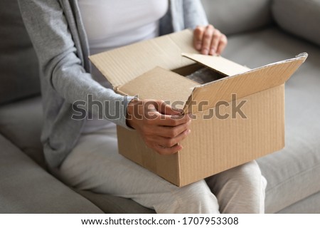 Close up woman holding parcel in hands, unpacking, curious young female opening cardboard box, gift, sitting on couch at home, client customer received online store order, delivery service Royalty-Free Stock Photo #1707953308