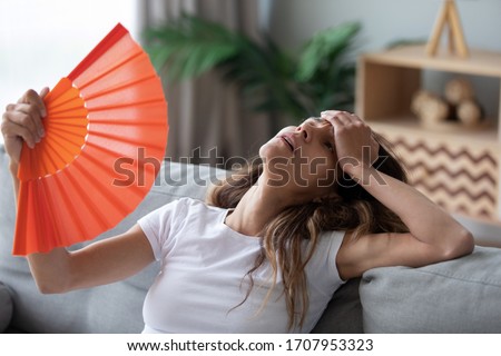Overheated woman sitting on couch, waving orange paper fan close up, girl feeling unwell, suffering from heating at home, feeling discomfort, hot summer weather or fever, sitting on couch alone Royalty-Free Stock Photo #1707953323