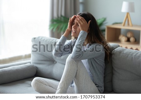 Stressed unhappy woman touching forehead, suffering from strong headache or chronic migraine, sick unhealthy girl feeling pain in head, sitting on couch at home alone, health problem Royalty-Free Stock Photo #1707953374