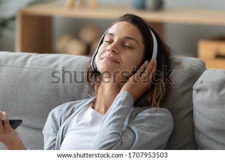 Satisfied woman in headset enjoying favorite music, relaxing on cozy sofa close up, happy young female wearing headphones listening to popular song, resting with closed eyes on soft pillow at home Royalty-Free Stock Photo #1707953503
