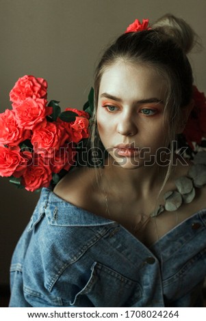 
Young beautiful woman with blond hair gathered in a bun (portrait). Sits in a denim blue jacket against a light brown wall. It is decorated with bright bushy red-pink roses with a eucalyptus branch.