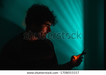 Man stays looking at his smartphone while an teal light penetrates the room with some shadow lines caused by the shutters of the window at his left