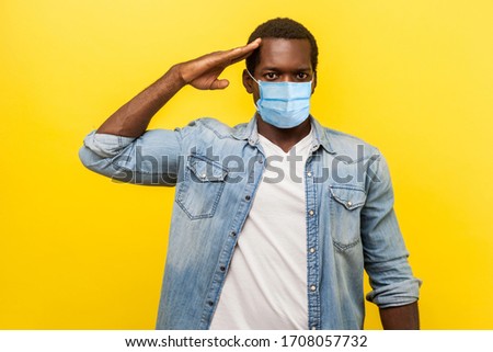 Yes sir! Portrait of serious confident man in denim casual shirt with surgical medical mask standing as soldier giving salute, ready for order. indoor studio shot isolated on yellow background