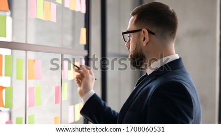 Serious middle-aged businessman in glasses brainstorm engaged in creative thinking in office, concentrated male employee write on colorful sticky notes develop business project at workplace Royalty-Free Stock Photo #1708060531
