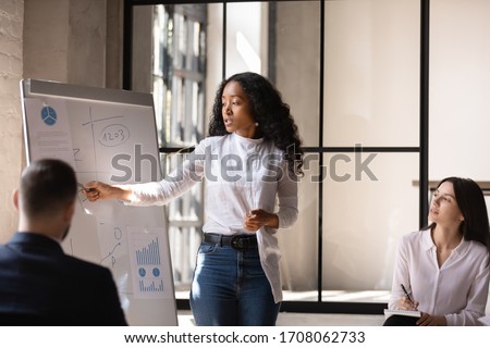 Young African American female coach or speaker make whiteboard presentation for diverse employees in office, focused biracial woman trainer present business project on flip chart at meeting Royalty-Free Stock Photo #1708062733