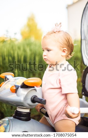 Lifestyle adorable cute blonde little baby girl plays on vacation hot summer day lit with sun rays. Child rides push along trike bicycle wearing pink princess crown.Happy childhood concept copy space
