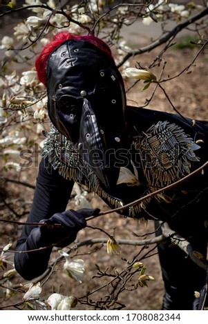 Young beautiful woman in a black leather bird mask with red hair in a black suit stands near a blossoming white magnolia in spring in the sunlight