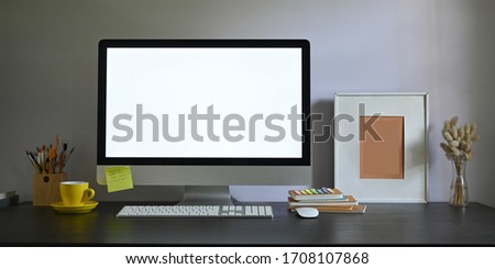 Photo of Workspace blank screen computer monitor putting on working desk and surrounded by picture frame, pencil holder, stack of books, wireless mouse, keyboard, coffee cup and wild grass in vase.