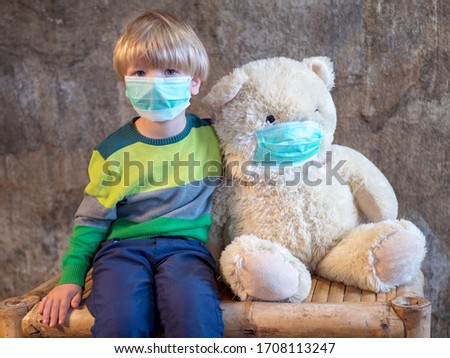 Funny boy in colored clothes plays in doctor and patient with white toy bear. child in blue medical mask put same on his friend. Toddler hugs and takes care of illness bear