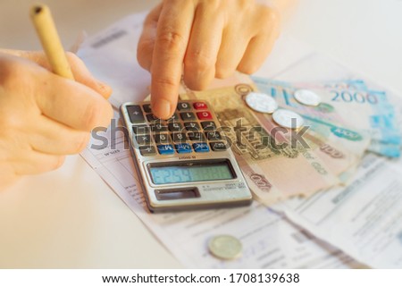 an elderly woman considers payments for utility bills; on a calculator, she considers payment for an apartment with money in Russian rubles; banknotes of 2000,100 rubles and Russian coins Royalty-Free Stock Photo #1708139638