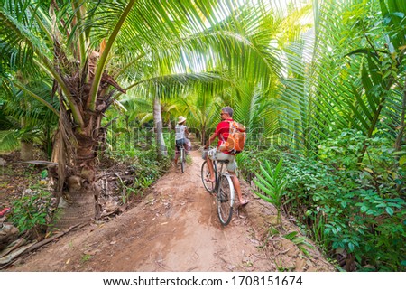 Tourist couple riding bicycle in the Mekong Delta region, Ben Tre, South Vietnam. Woman and man having fun cycling on trail among green tropical woodland and coconut palm trees. Rear view.
