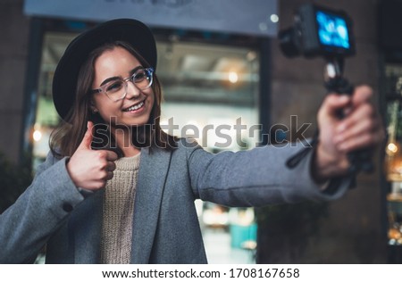 Female vlogger recording with digital camera showing ok or like. Smiling woman taking selfie video on light night city. Traveler making video for her blog. Vlogger uses photo camera for shoot social