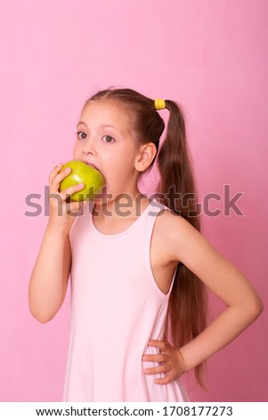 Studio close-up portrait of caucasian  girl with funny hairstyle, with eyes widened, biting big green apple, over pink background