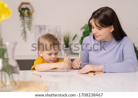 Pretty young mother and her cute little son drawing pictures with colorful crayons while staying at home during period of quarantine
