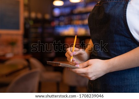 Hands of young waitress in dark blue apron holding pencil over page of notepad while going to write down order of client in restaurant Royalty-Free Stock Photo #1708239046