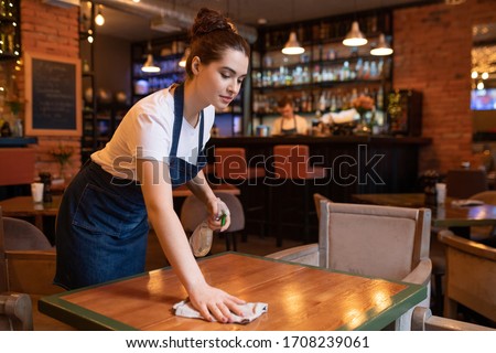 Pretty young waitress in apron bending over wooden table while using detergent and duster to clean it for new guests of restaurant Royalty-Free Stock Photo #1708239061