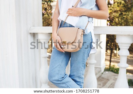 Bag in women's hands. Details of casual summer or spring outfit. Woman wearing blue jeans, white t shirt, small beige cross body bag standing outdoors. Everyday look. Street fashion. No face. Royalty-Free Stock Photo #1708251778