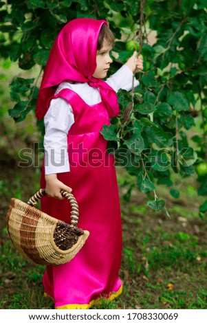 kid girl in a pink shawl and dress like Masha and the bear from the cartoon holds a wicker basket and picks apples.