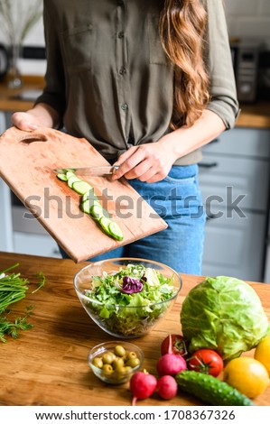 Healthy and easy food concept. Close-up picture. Hands of woman throw sliced cucumber from cutting board in a bowl of a salad. Wooden table with vegetable on it