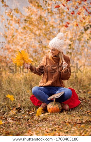 female portrait. Cute smiling blonde in a white knitted hat, a brown sweater sits on a plaid in the park, holds a maple leaf and looks into the distance. vertical photo