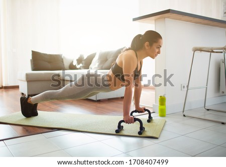 Young woman doing sport workout in room during quarantine. Picture of strong fitness model stand in plank position using push up stands hand bar. Exercising in room alone.