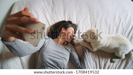 Authentic shot of an young happy woman is making a selfie or video call to a boyfriend with her puppy of Labrador Retriever dog in a bed. Concept of technology, connection,pets, family authenticity
