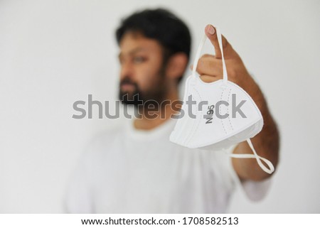 Indian man holds up a white N95 mask in clear focus while the person holding it is out of focus. Shallow depth of field, mask for protection against corona virus, Omicron and other airborne diseases Royalty-Free Stock Photo #1708582513