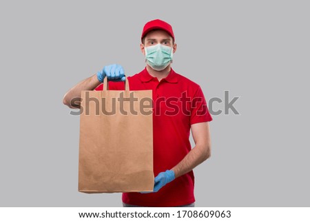Delivery Man Wearing Medical Mask and Gloves With Paper Bag in Hands. Red Uniform Delivery Boy. Home Food Delivery. Paper Bag Royalty-Free Stock Photo #1708609063