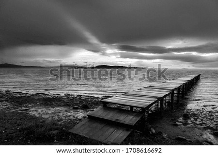 Low point view of a pier on a lake with a ray of sun against a moody sky