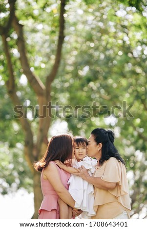 Beautiful granny and mother hugging and kissing little girl when standing in park