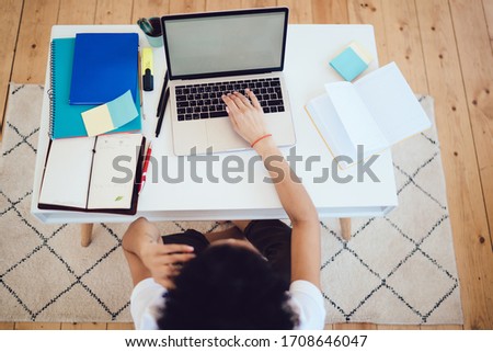 From above unrecognizable young female freelancer in casual wear having phone conversation while working with laptop and sitting on floor at home 
