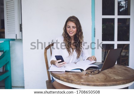 Happy charming blond business woman in white shirt flipping book pages and using smartphone while sitting at rounded table with laptop outdoors