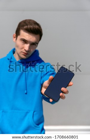 Serious slender young shaved man in a blue hoodie look in to the cam and holds out his smartphone with six inch display in his left hand, standing against a gray textured wall in the afternoon